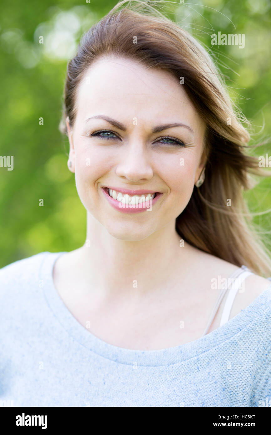 Porträt einer schönen Frau. Sie steht im Freien mit ihrem Haar im Wind und Bäume sind unscharf im Hintergrund. Stockfoto