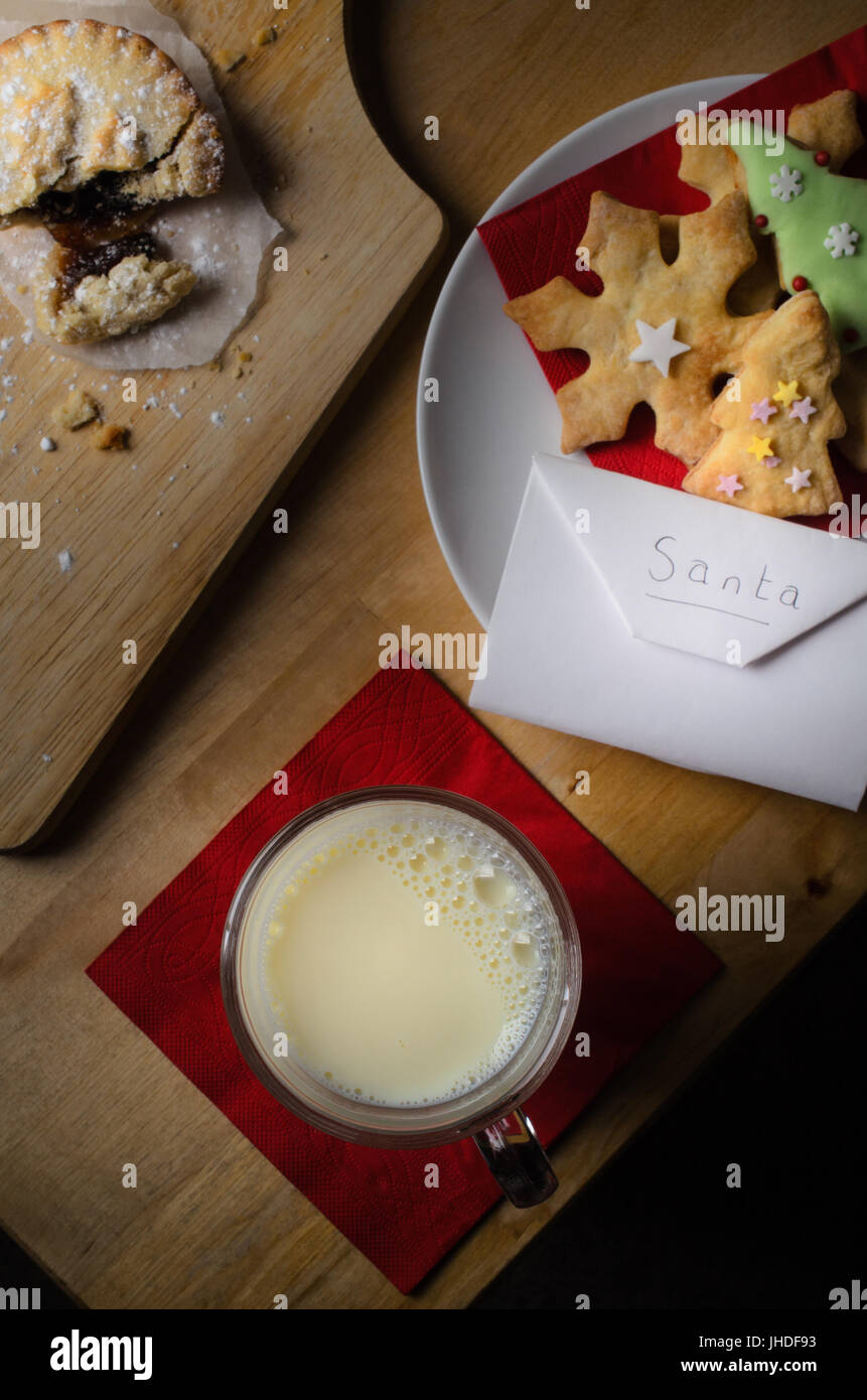 Kekse (Cookies), Milch und Mince Pie ausgelassen auf Holztisch für Santa Claus mit Umschlag mit Wunschliste.  Mince Pie ist wie du aufgebrochen Stockfoto