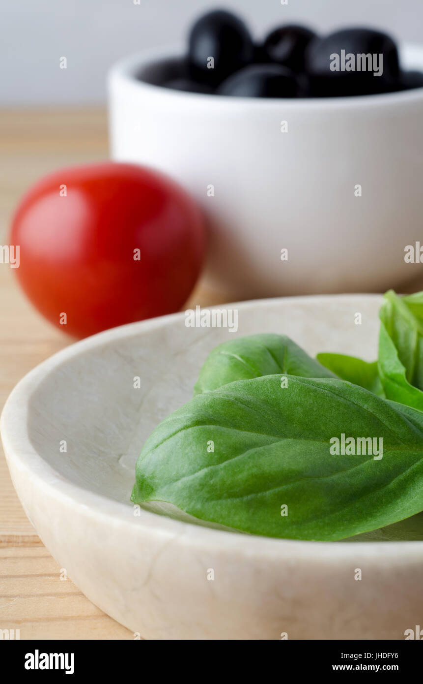 Servierplatte von frisch gepflückten grünen Basilikum-Blätter im Vordergrund mit roten Tomaten und schwarzen Oliven in weiße Schüssel im Hintergrund. Stockfoto