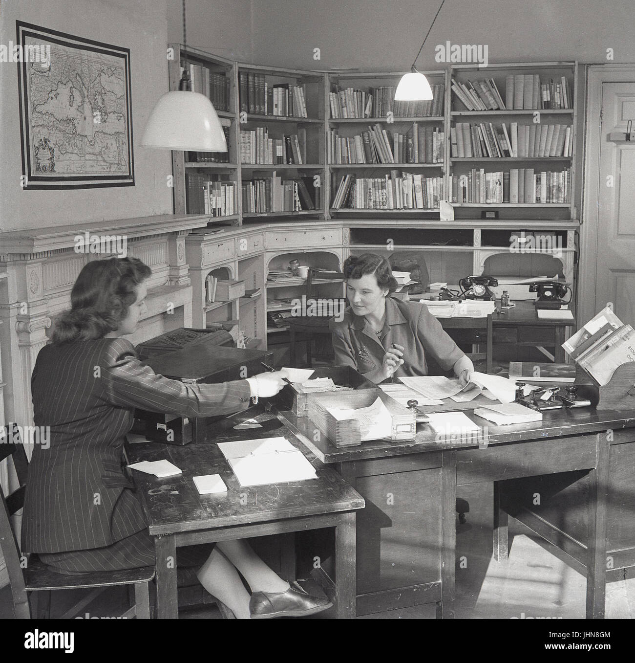 1950, historische, England, UK, Bild zeigt einen weiblichen Leiter einer Hochschule an ihrem Schreibtisch arbeiten in ihrem Buch Büro gefüllt, mit einem intelligenten weiblichen Assistenten helfen bei der Büroarbeit und Verwaltung. Stockfoto