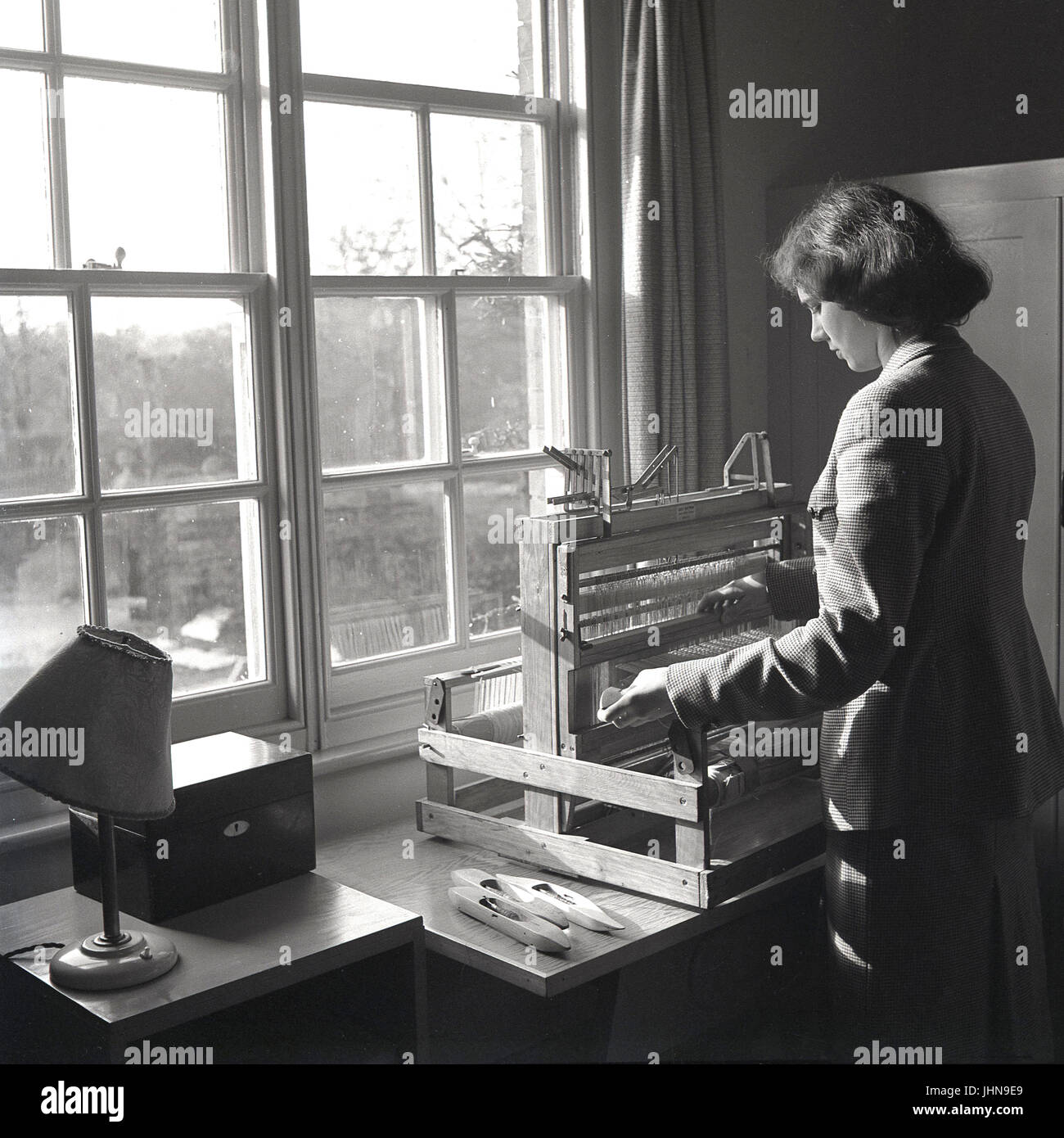 1950, historische, Bild zeigt eine junge Frau durch ein Schiebefenster mit einem traditionellen kleinen Holztisch - top Hand-Webmaschine oder Webstuhl tp ein Stück Stoff oder Stoff produzieren, England, UK. Stockfoto