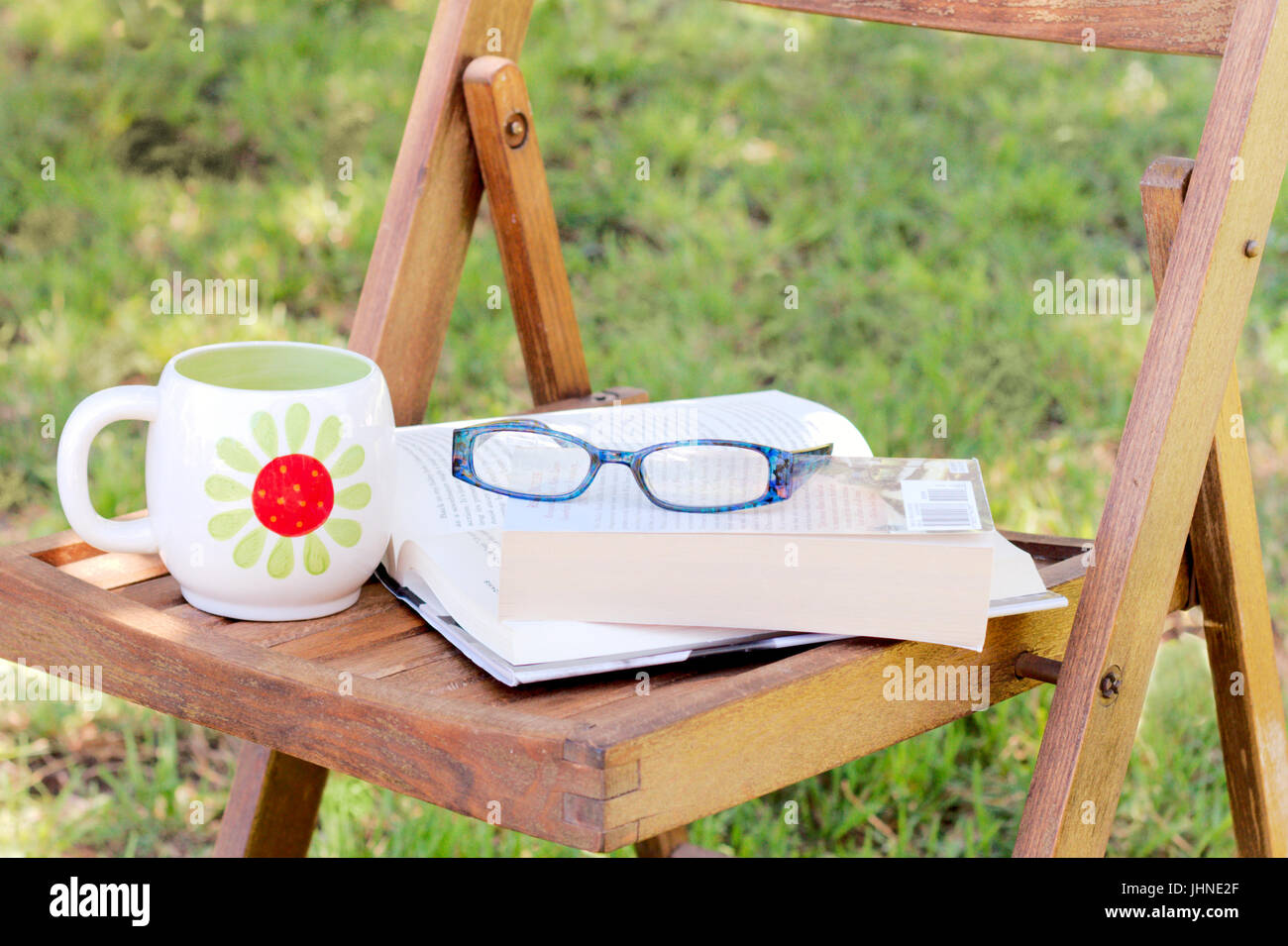 Bücher mit Lesebrille und Tasse Tee ruhen auf einem Holzstuhl im gefleckten Schatten. Stockfoto