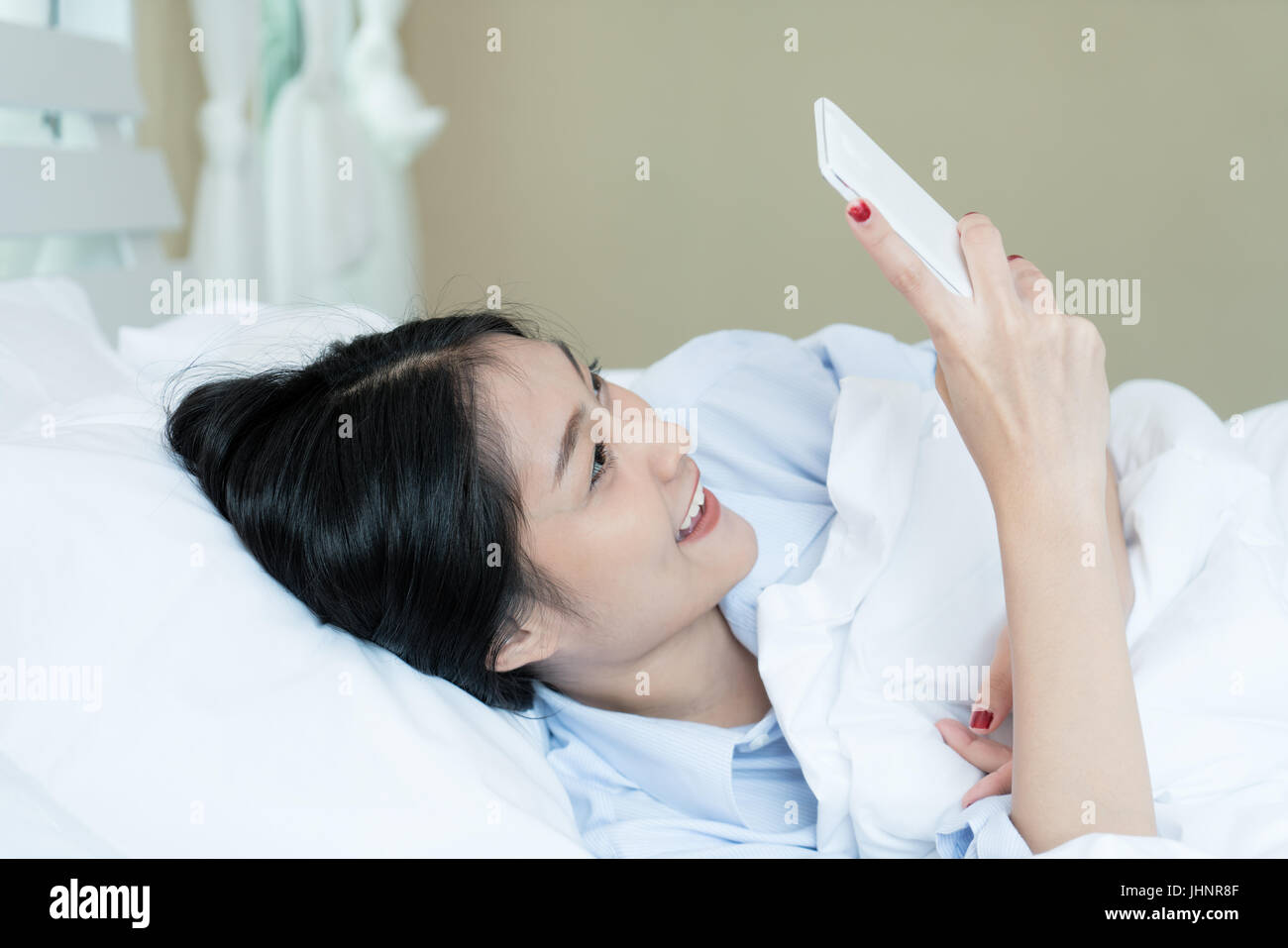 Schöne asiatische Teenager Frau im weißen Bett liegend und mit einem Telefon in ihrem Schlafzimmer zu Hause. Technologie im Wohnkonzept. Stockfoto