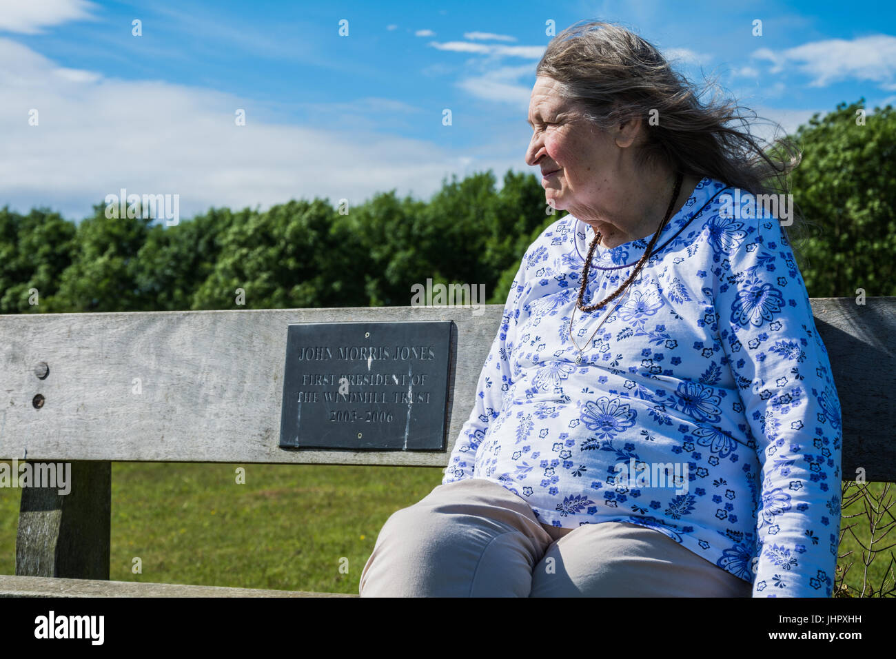 Ältere Frau auf einer Bank sitzend genießen ihren Ruhestand an einem sonnigen Tag mit einer Sommerbrise weht durch ihr Haar. Stockfoto