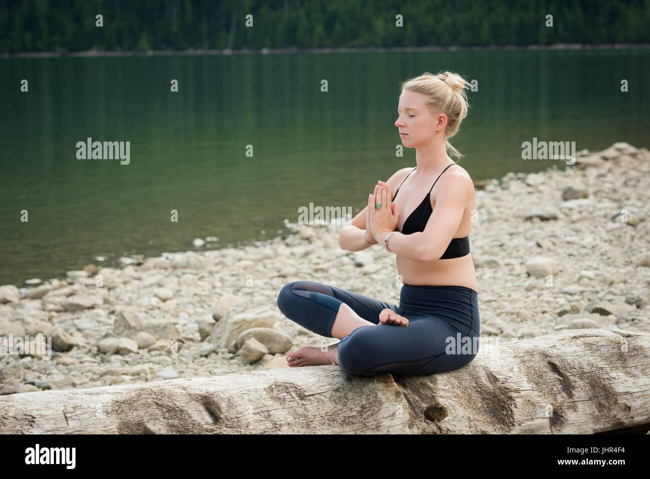 Junge Frau beim Üben von Yoga auf Log am Seeufer zu meditieren Stockfoto