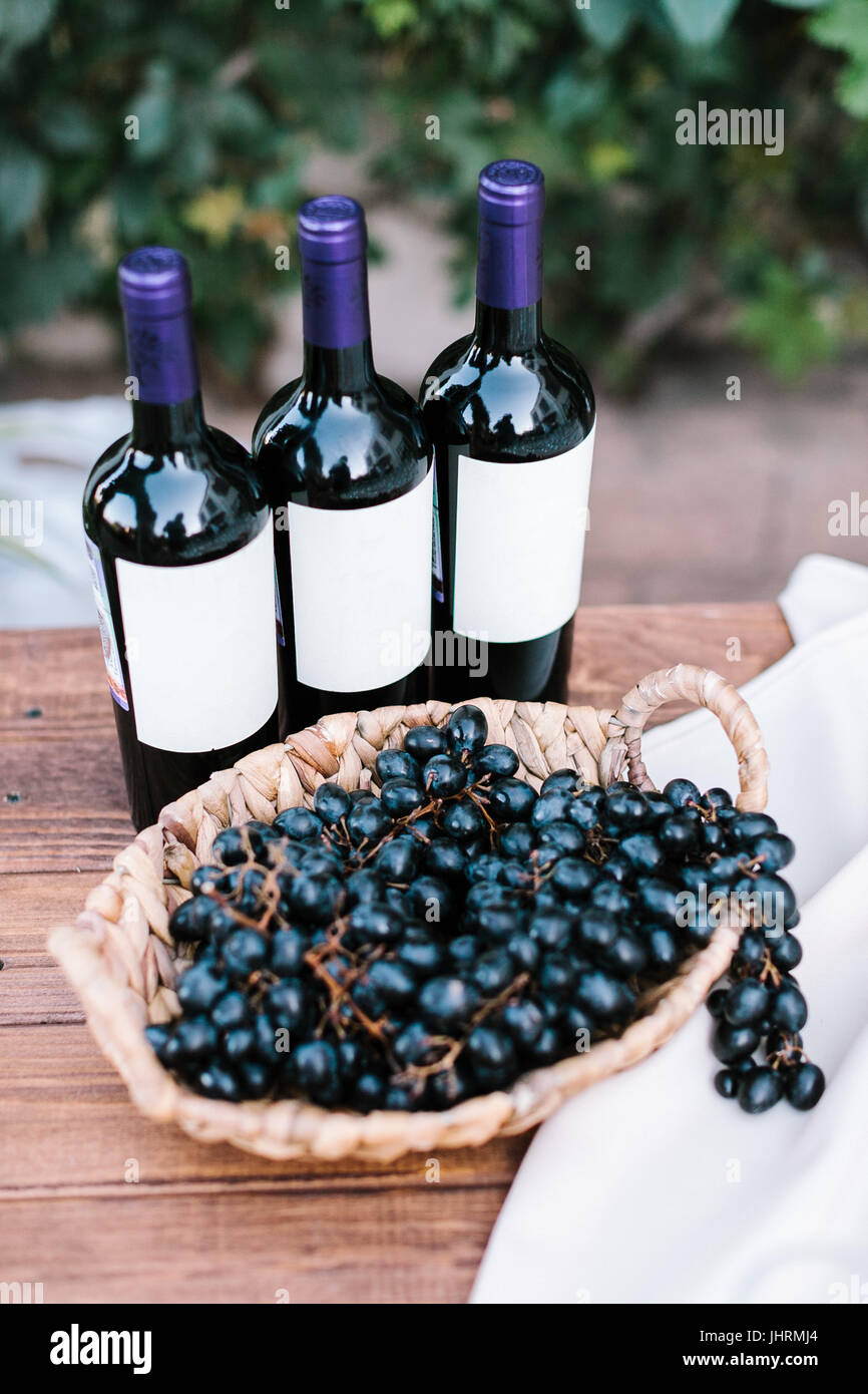 drei Flaschen Rotwein und Ansammlung von Labruscan Trauben in einer wattled Platte auf einem Holztisch im Freien. Aperitif an einer Hochzeit. Stockfoto