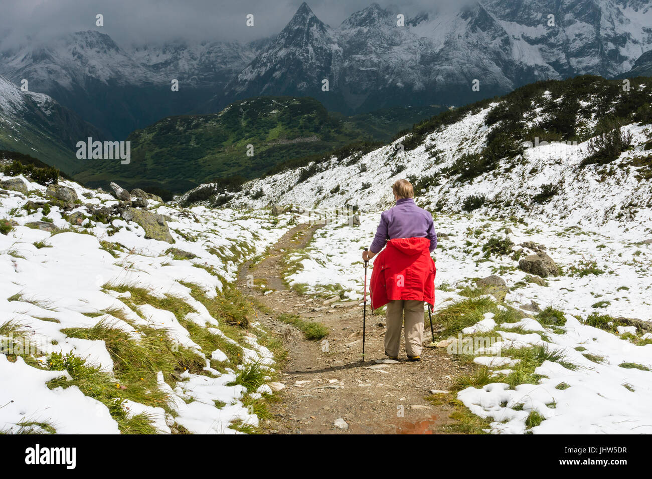 Ein weiblicher senior Wanderer trägt einen roten Mantel, Wandern durch Schnee im Sommer auf dem Weg zum Zeinisjoch in das Paznaun-Tal in der Nähe von Galtur in Österreich. Stockfoto