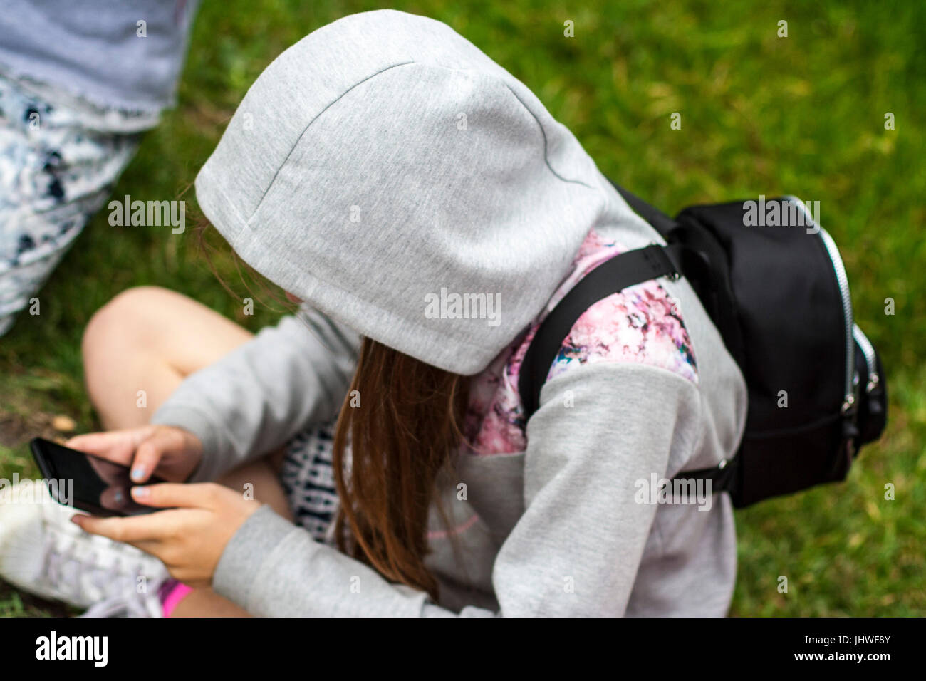 Teenager sitzen gekreuzten Beinen auf Gras mit Kapuze bis auf Ihr Telefon im Sommer suchen, Dublin Irland jugendlich Mädchen Stockfoto