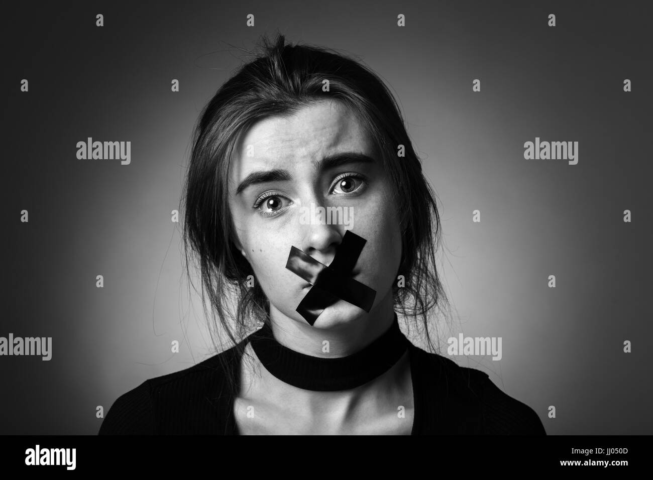 Trauriges Mädchen mit Klebeband auf den Mund, der Blick in die Kamera auf grauem Hintergrund, Monochrom Stockfoto
