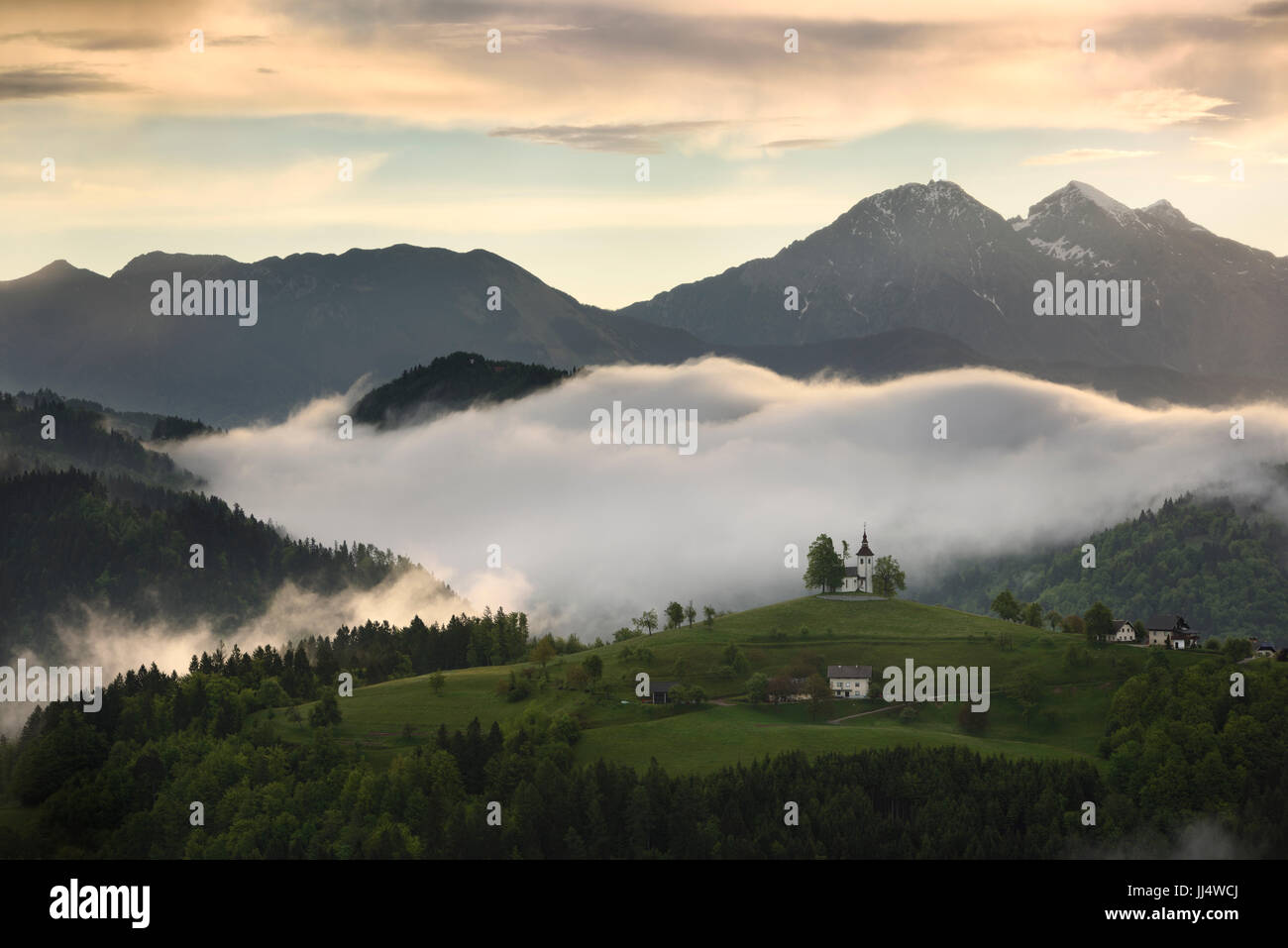 Rollende Nebel bei Sonnenaufgang in den Skofjelosko Hügeln mit St. Thomas Kirche mit Kamnik Savinja Alpen in der Nähe von Ljubljana Slowenien Stockfoto