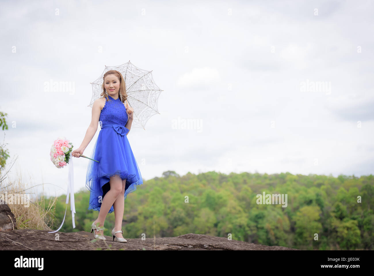 Schöne Frau tragen blaue Abendkleid halten einen Strauß Blumen und weißen Schirm. über Berge und Flüsse Hintergrund. Stockfoto