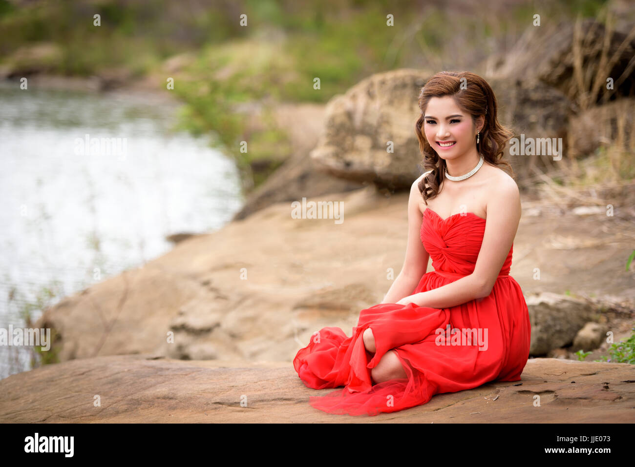 Schöne Frau tragen rote Abendkleid glückliches Lächeln sitzt auf einem Felsen im Wald hautnah. Stockfoto