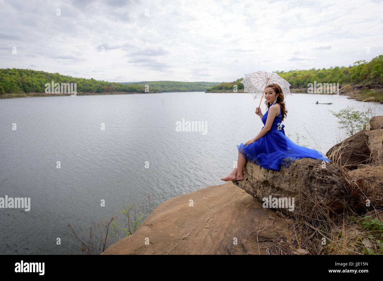 Junge Frau trägt blaue Kleider halten einen weißen Schirm beim Blick auf den Fluss und die Berge. Stockfoto