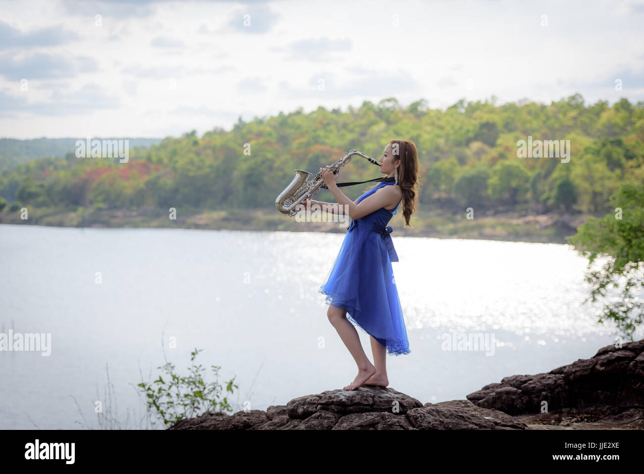 Schöne Frau tragen blaue Abendkleid sound Saxophon Ständer auf den Felsen über Berge und Flüsse Hintergrund. Stockfoto