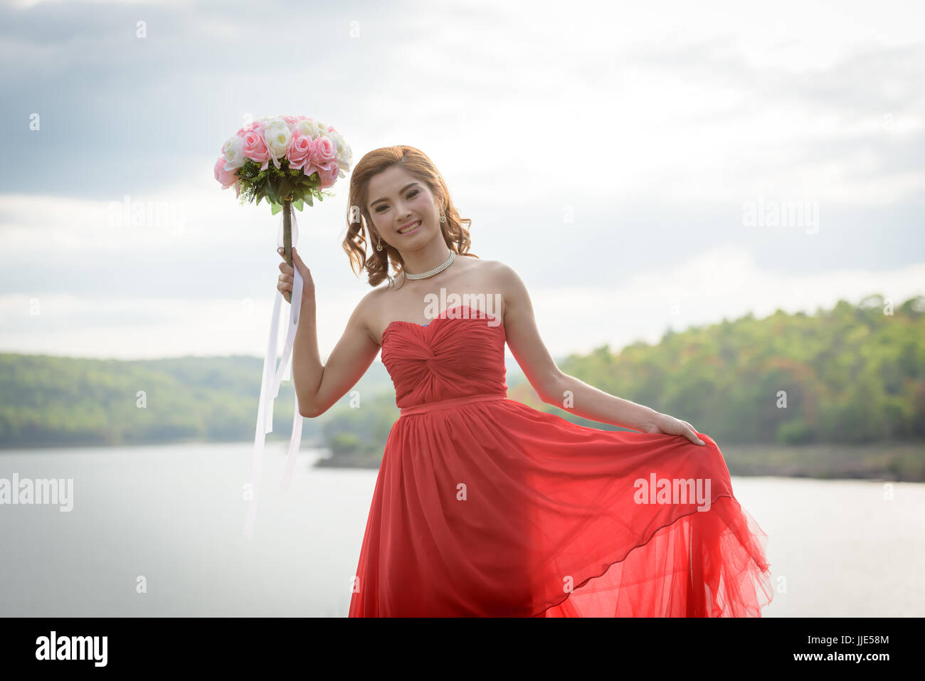 Schöne Frau tragen rote Abendkleid halten einen Blumenstrauß. über Berge und Flüsse Hintergrund. Stockfoto