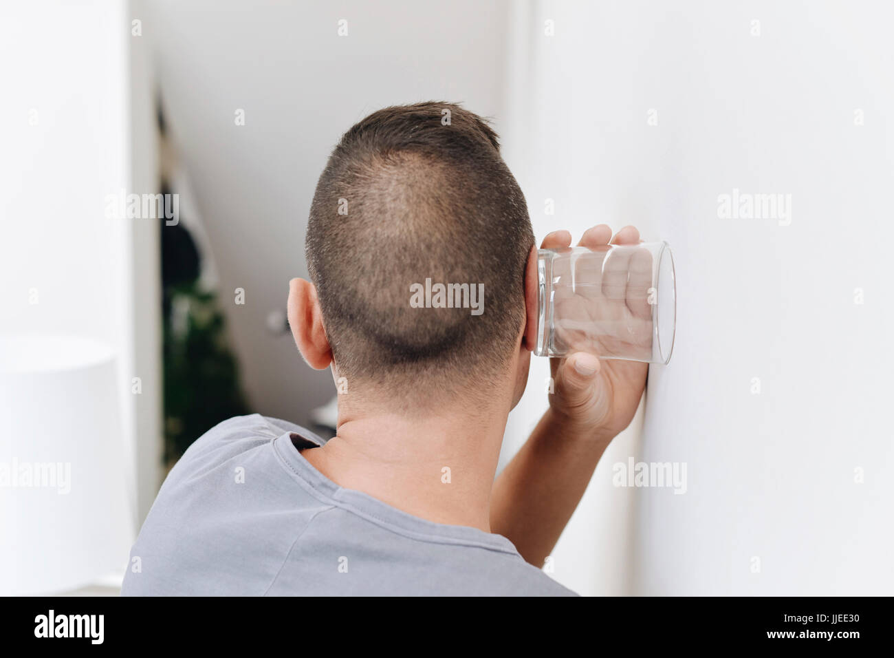 Closeup von einem kaukasischen Jüngling gesehen von hinten durch die Wand mit einem Glas hören wollen Stockfoto