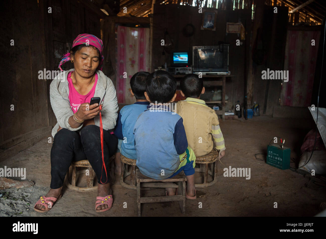 Eine Mutter spielt Musik von ihrem Handy, während ihr Sohn und zwei seiner Freunde Fernsehen zu Hause in Ban Sop-Kha, Laos. Die Elektronik werden von Batterien (rechts) durch Mikro-hydro Turbinen behelfsmäßig in den Nam Ou Fluss angetrieben. Stockfoto