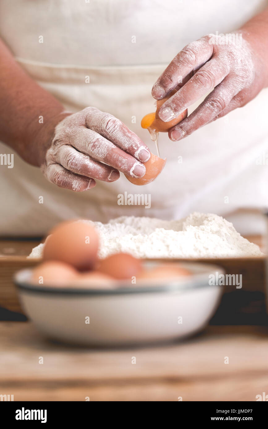 Ältere Frau Teig Ei hinzufügen. Fokus auf gebrochene Ei und alte arbeitende Hände. Vertikaler Ausrichtung, gesättigte verblasst Farben Stockfoto