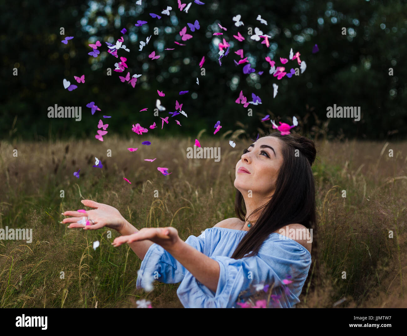 Elfenbein, rosa und lila farbige Schmetterling Konfetti-Regen nach unten auf eine schöne junge Frau während einer Sommer-Porträt-Sitzung auf einer Wiese in der Nähe von Liverpool. Stockfoto