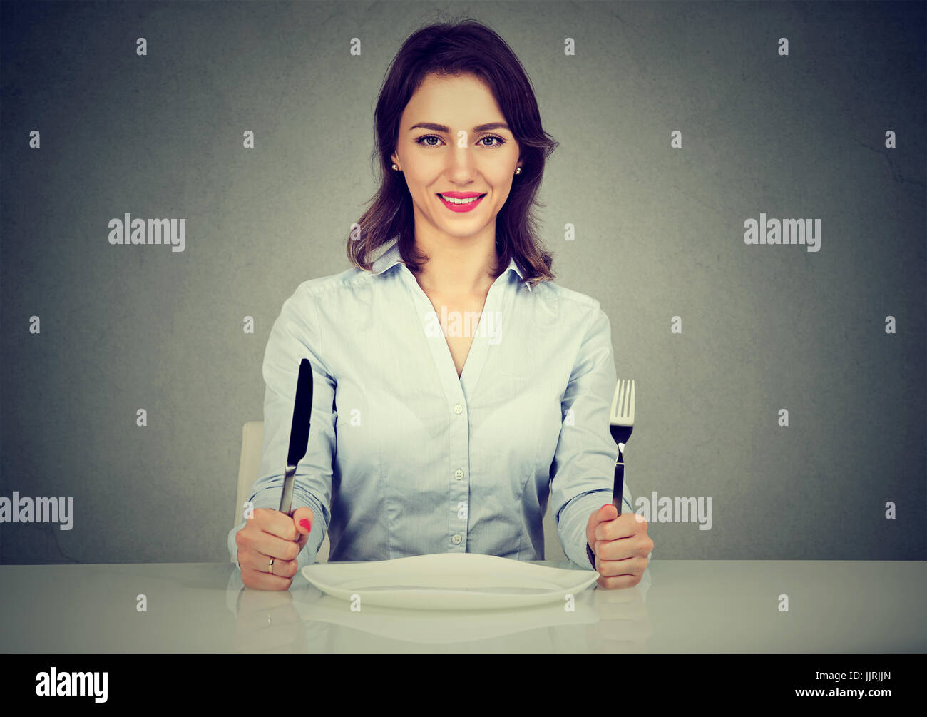 Glückliche Frau mit Gabel und Messer mit leeren Teller auf graue Wand Hintergrund isoliert am Tisch sitzen Stockfoto