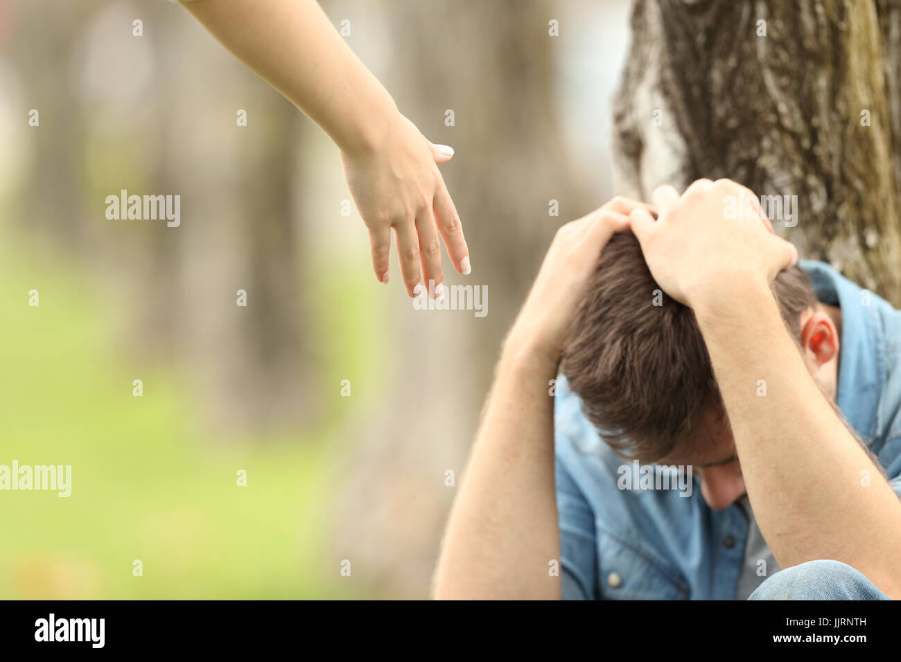 Traurige Teenager sitzen auf dem Rasen in einem Park und die Hand einer Frau bietet Hilfe mit einem grünen Hintergrund Stockfoto
