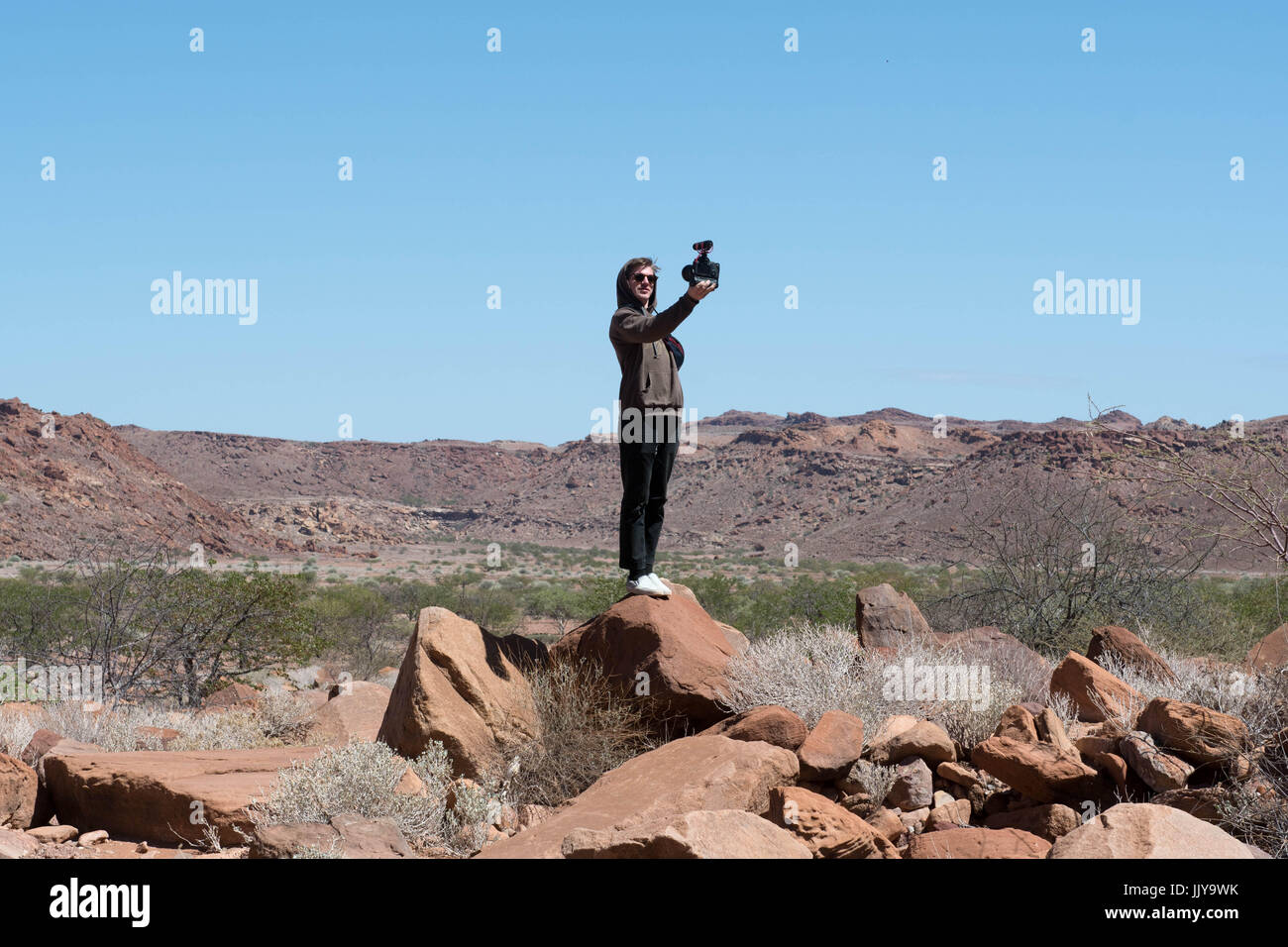 Ein Tourist steht auf einem Felsen und nimmt ein Selbstporträt in Twyfelfontein, dem Gelände der alten Steinmetzarbeiten von alten Damaraland Stämme lo gemacht Stockfoto