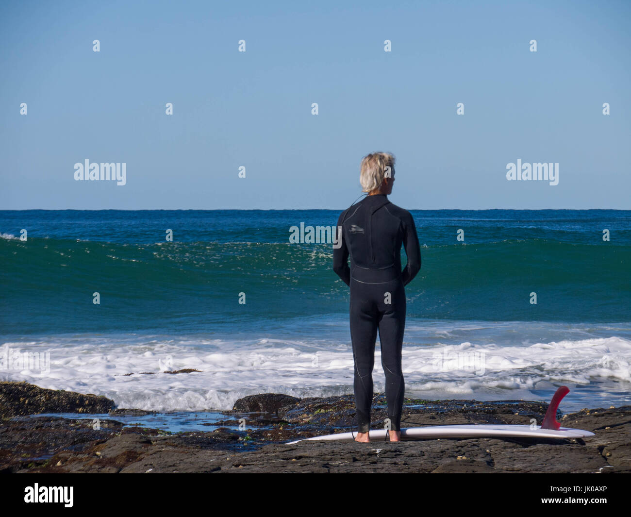 Eine ältere männliche Surfer stehen und warten auf den Wellen Shorebreak mit seinem Surfbrett auf den Felsen am Meer Stockfoto