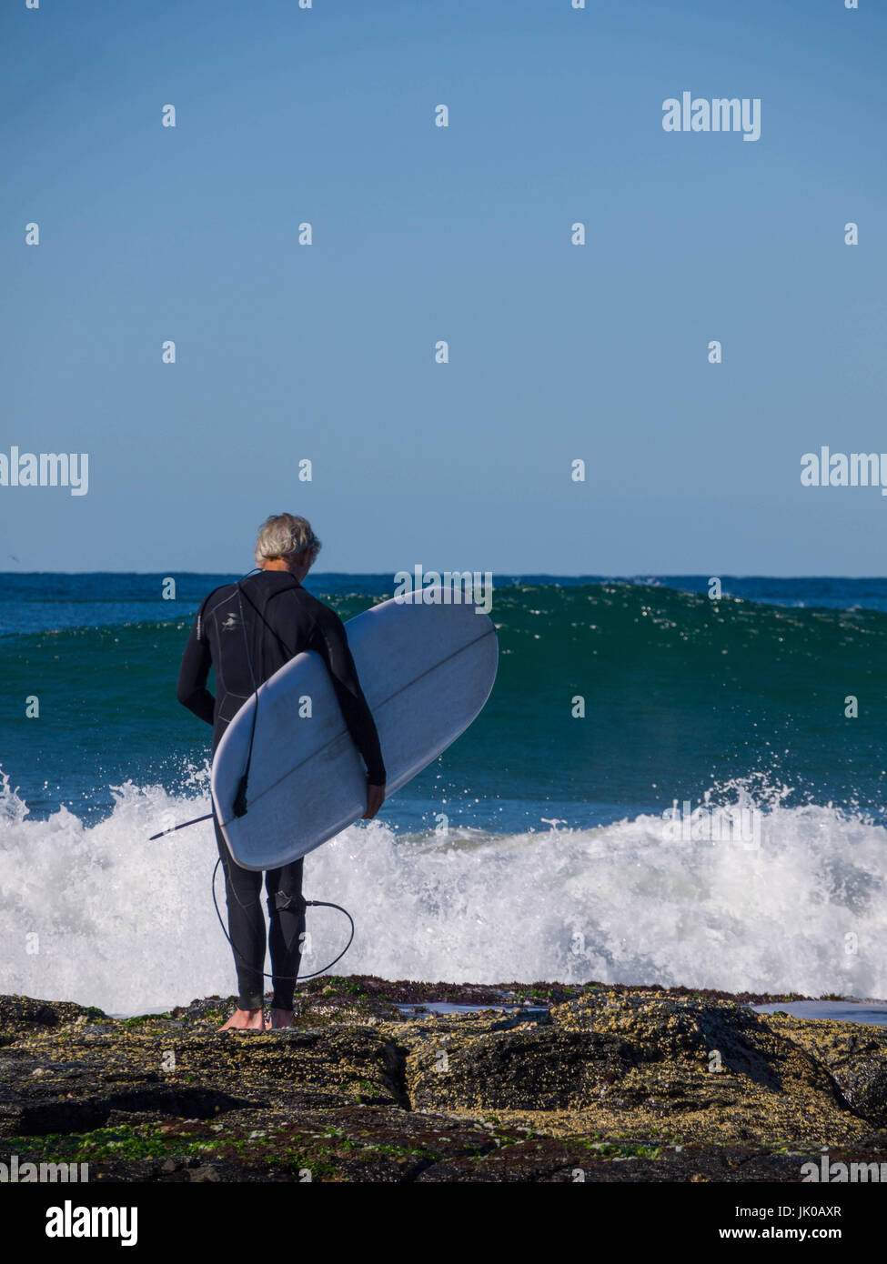 Eine ältere männliche Surfer stehen und warten auf den Wellen Shorebreak mit seinem Surfbrett auf den Felsen am Meer Stockfoto