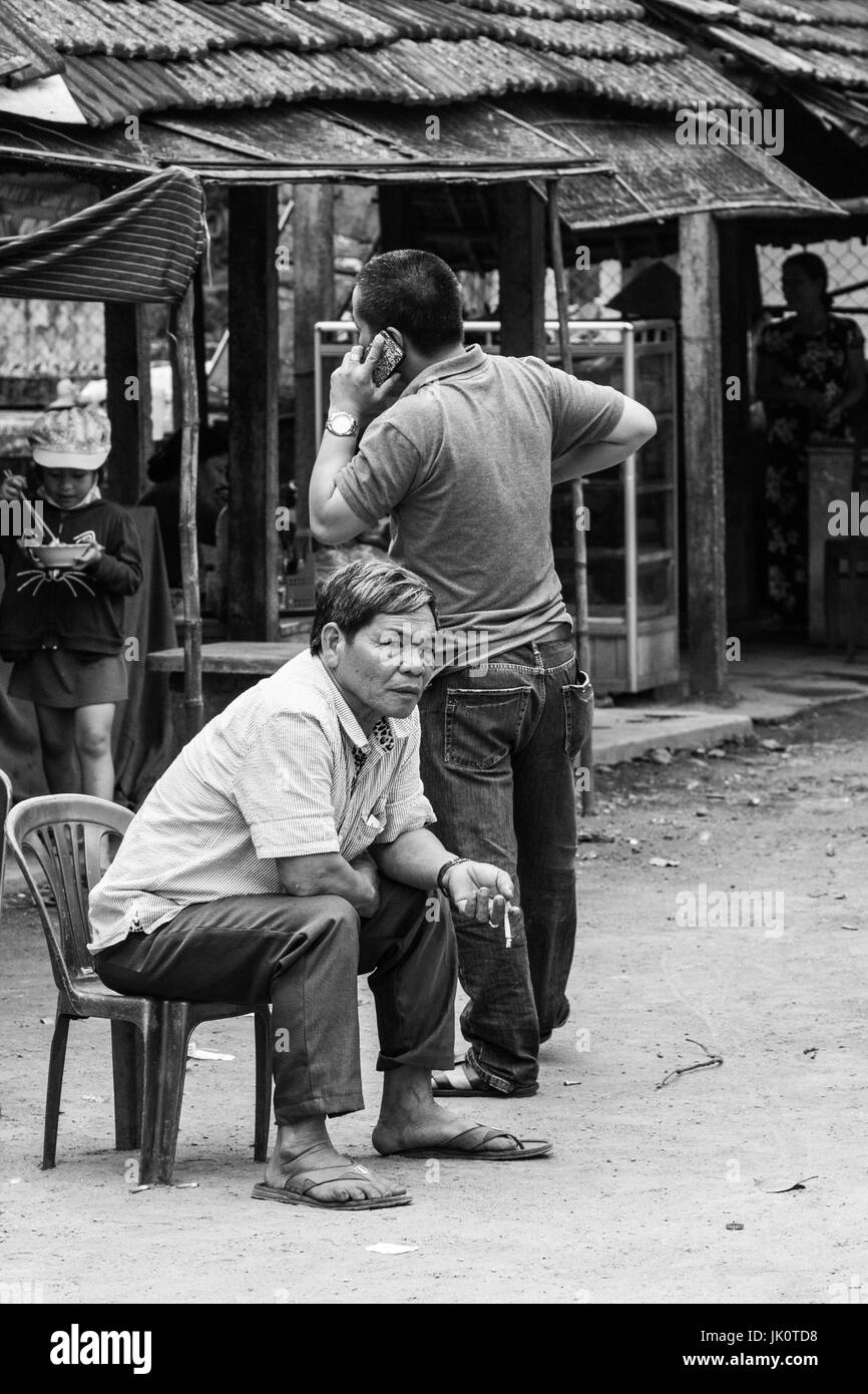Ein Mann sitzt in Gedanken verloren und vergessen, den chaotischen Markt um ihn herum, wie er eine Zigarette raucht Stockfoto