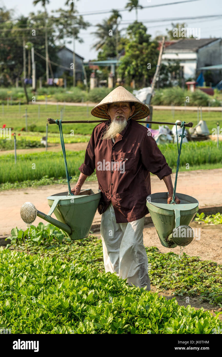Vietnamesische Greis in seinen Neunzigern Bewässerung von Pflanzen mit schweren Gießkannen Stockfoto
