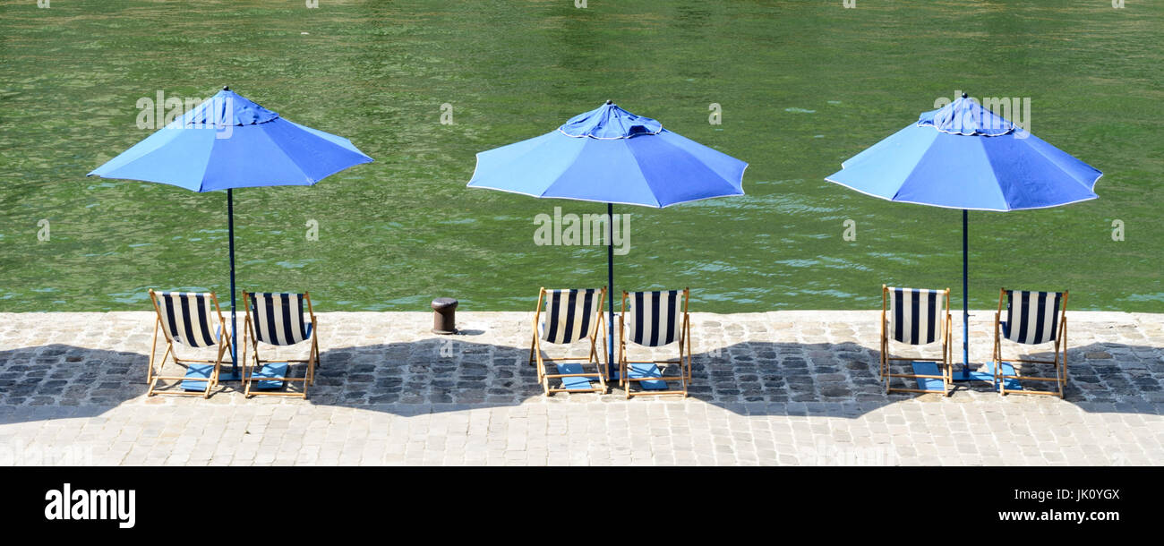 Drei blaue Sonnenschirme mit blau-weiß gestreiften Liegestühlen in der Sonne am Kai des Flusses Seine mit grünem Wasser. Stockfoto