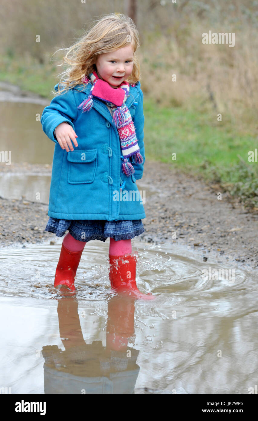 Mädchen in roten Gummistiefeln und Blue Coat watet durch eine Pfütze Stockfoto