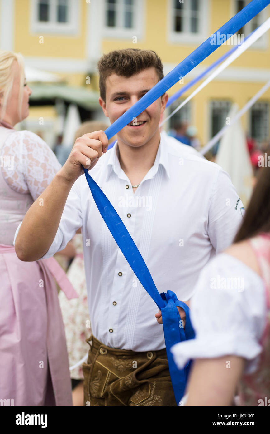Bayerische junge Mann im weißen Hemd und Lederhosen hält blaues Band während der Durchführung traditionellen Volkstanz Bandltanz rund um den Maibaum Stockfoto