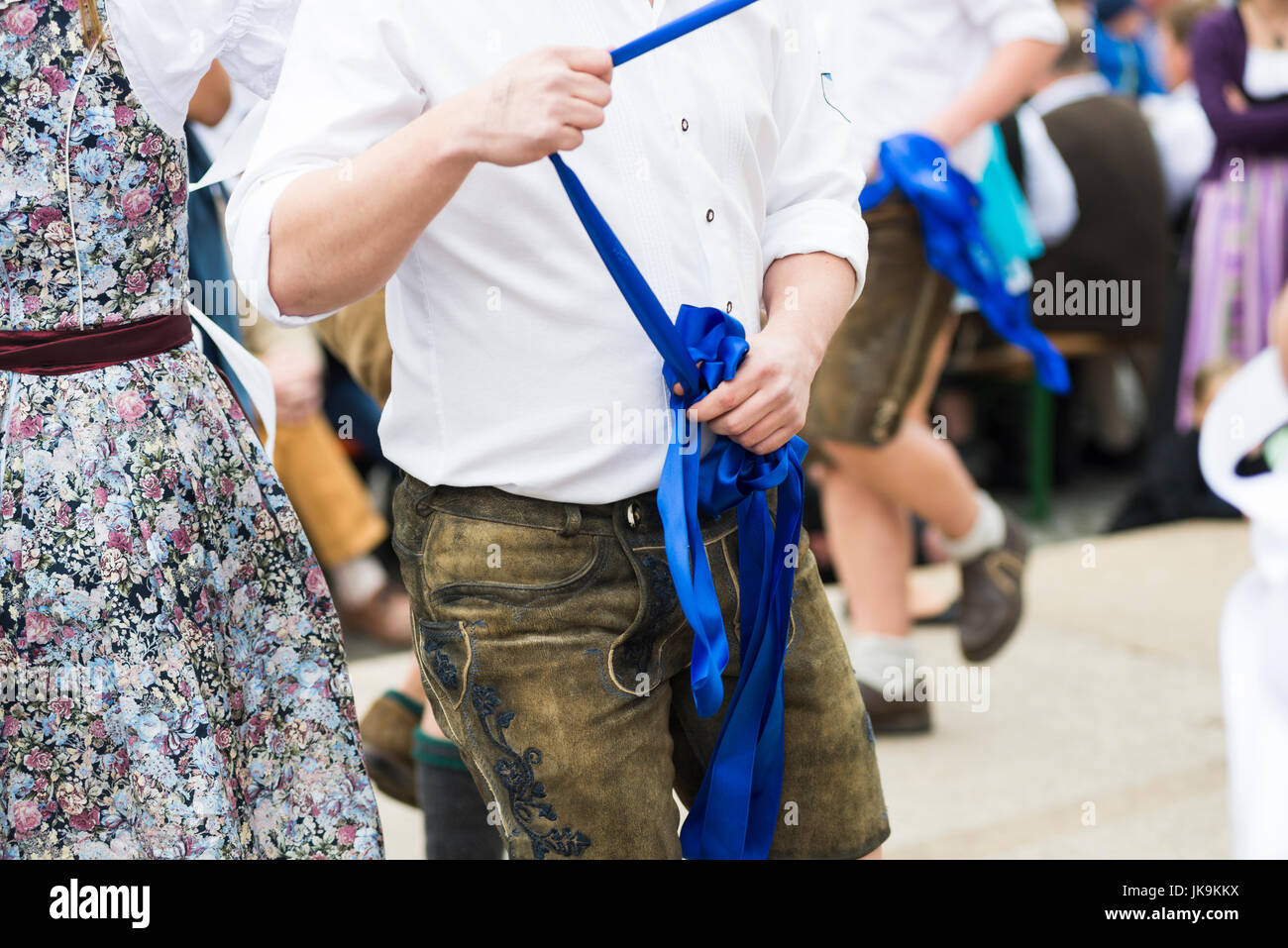 Junger Mann im weißen Hemd mit Lederhose hält blaues Band beim Tanzen um den Maibaum Stockfoto