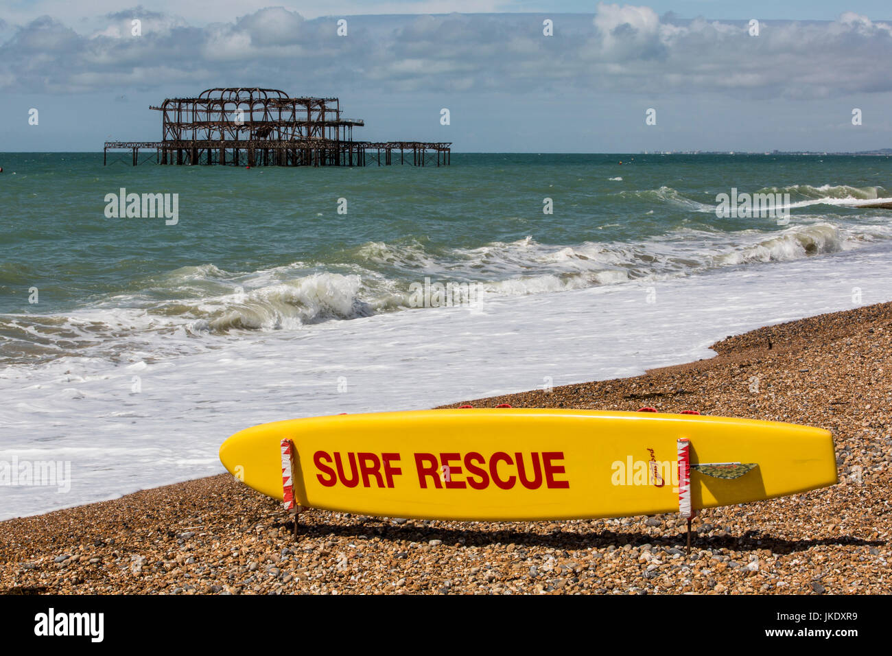 Surfbrett mit 'Surf Rescue"gemalt, auf einem Kiesstrand am Meer, mit die ausgebrannten Reste der Brighton Pier hinter. Stockfoto