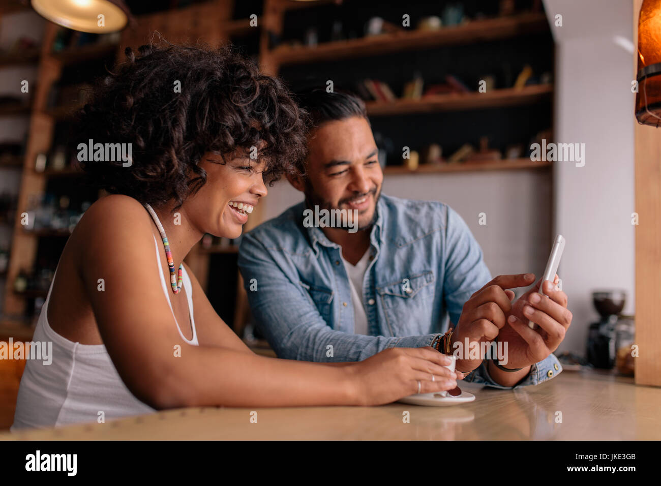Junge glückliche Paar am Tisch im Café sitzen und mit Blick auf Handy und lächelnd. Afrikanerin und kaukasischen Mann in Coffee-Shop mit Smartphone. Stockfoto