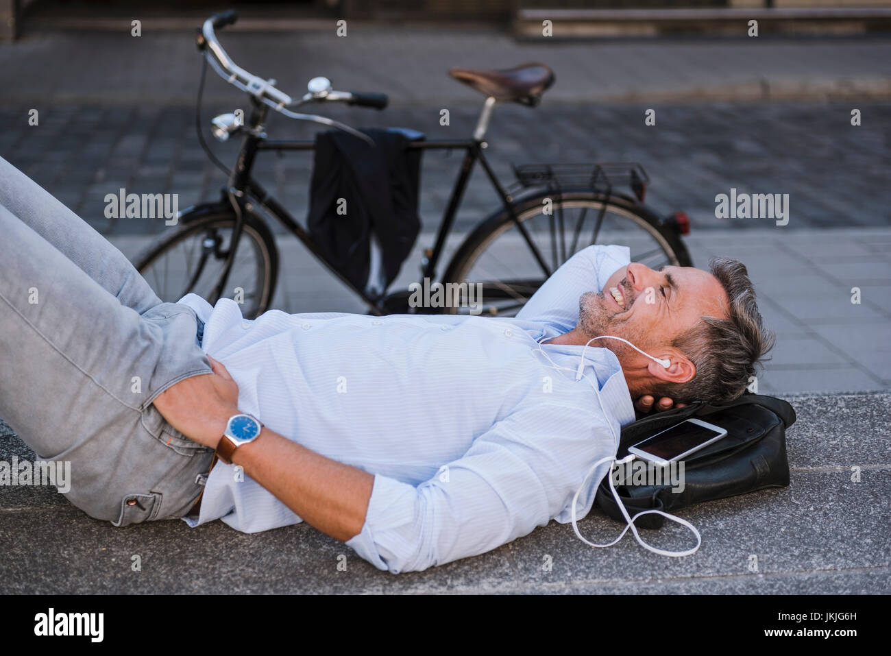 Lächelnder Mann mit geschlossenen Augen liegen auf Treppen mit Handy und Ohrhörer Neben fahrrad Stockfoto