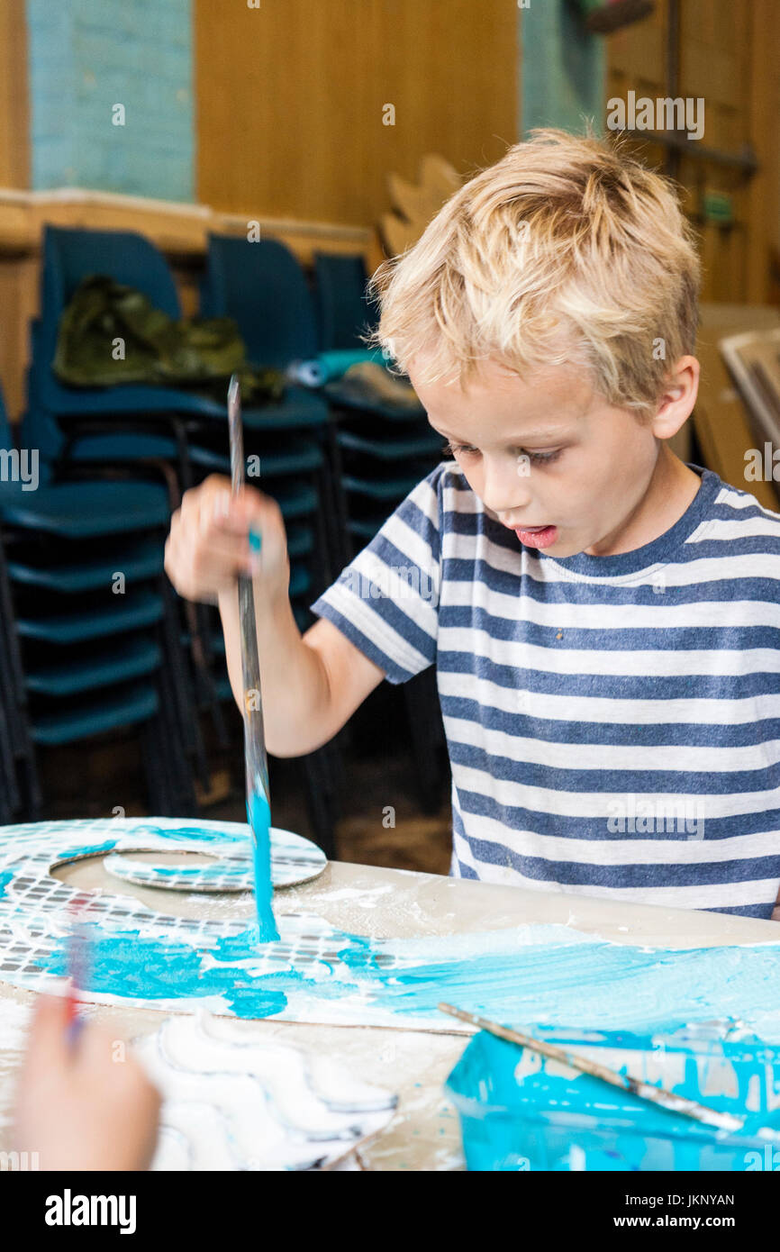 Kaukasische blonde Kind, Junge, 6-7 Jahre alt, Malerei der blauen Farbe auf die Pappe aus scharfen Schnitt. Drinnen in Kunst und pädagogische Werkstatt Handwerk. Stockfoto