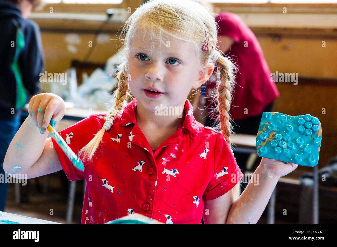 Kaukasische blonde Kind, Mädchen, 5-6 Jahre alt, blau Stück gemalte Karte und in der anderen Hand einen Pinsel. Kopf und Schultern. Zeigt. Drinnen in Kunst und pädagogische Werkstatt Handwerk. Stockfoto