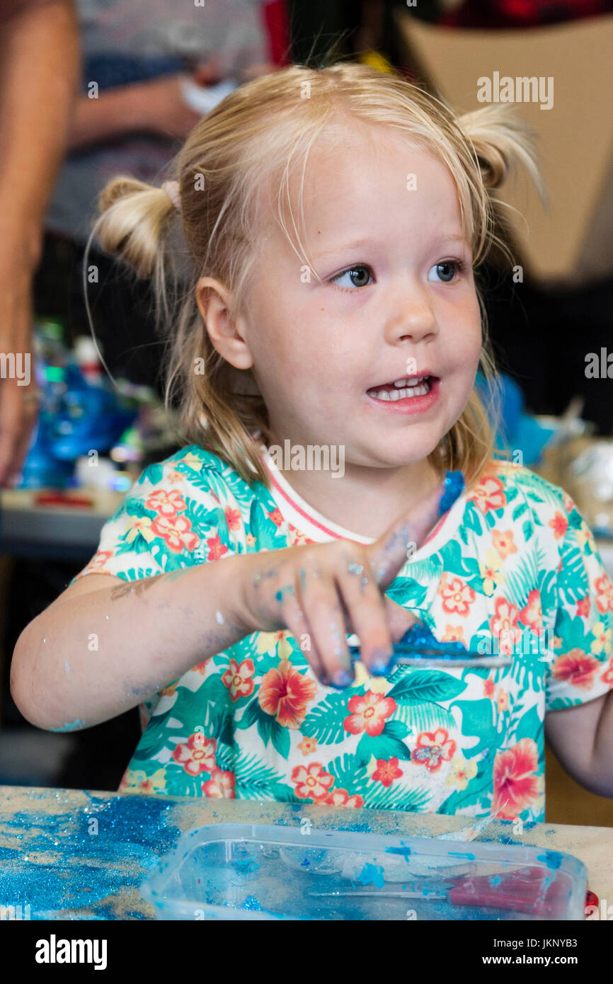 Kaukasische blonde Kind, Mädchen, 5-6 Jahre alt, Ihren Finger Anzeige mit blauer Farbe auf. Blick, leicht weg schauen. Hallenbad in Kunst und Handwerk Lernwerkstatt. Stockfoto