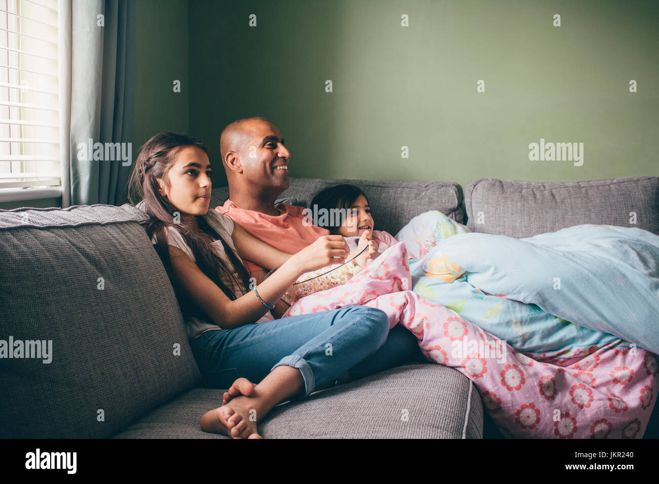 Vater zusammengerollt auf dem Sofa mit seinen beiden Töchtern. Sie befinden sich gerade einen Film mit Popcorn. Stockfoto
