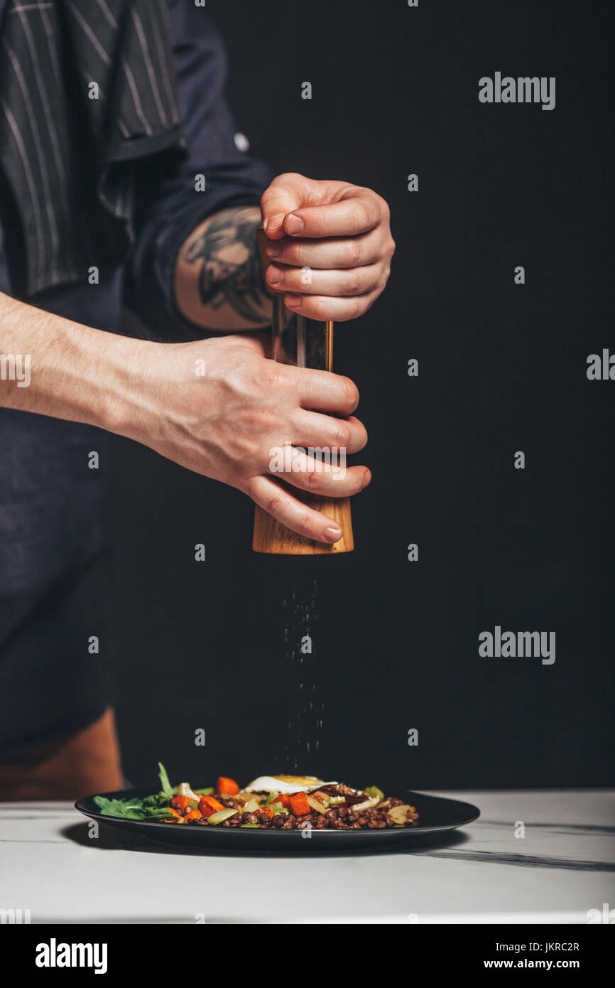 Beschnitten, Menschenbild, die Beregnung von Pfeffermühle über Essen auf Tisch vor schwarzem Hintergrund Stockfoto