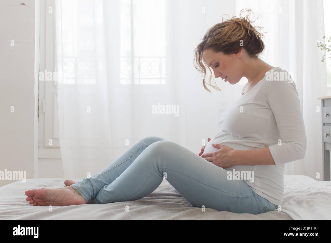 Schwangere Frau auf dem Bett sieht am Bauch sitzen Stockfoto Schwangere Frau auf dem Bett sieht am Bauch sitzen Stockfoto