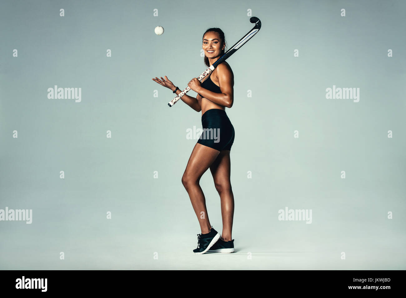 In voller Länge Portrait glückliche junge Frau mit Hockeyschläger und Ball über grauen Hintergrund. Hispanic weibliche Eishockeyspieler, lächelnd. Stockfoto