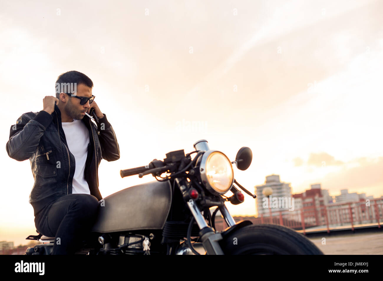 Hübscher Reiter Mann mit Bart und Schnurrbart Rauchen Strafpredigt und richtige Biker-Jacke sitzen auf klassischen Stil Café Racer Motorrad bei Sonnenuntergang. Fahrrad benutzerdefinierte m Stockfoto