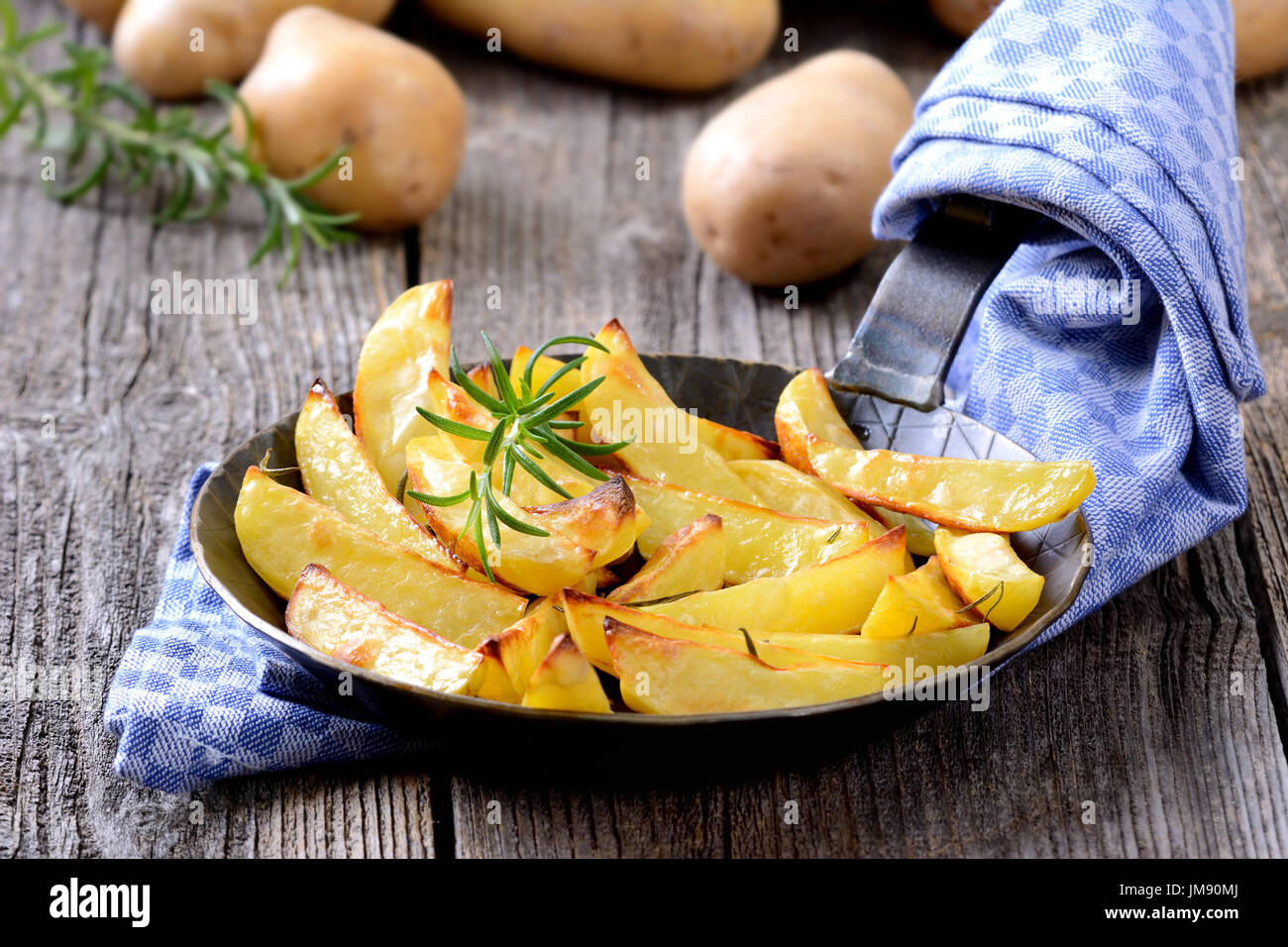 Gebackene Kartoffelspalten mit Rosmarin in einer Eisenpfanne, rohen Kartoffeln im Hintergrund Stockfoto