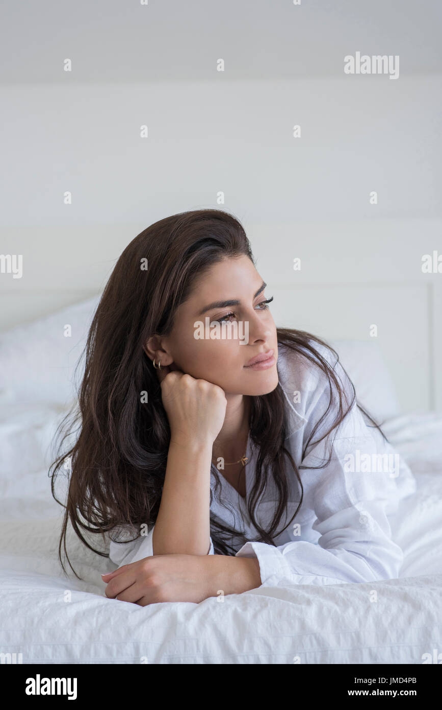 Junge Frau am Bett liegen und trägt ein weißes Hemd. Sie stützte sich auf Ellbogen und wegsehen. Stockfoto