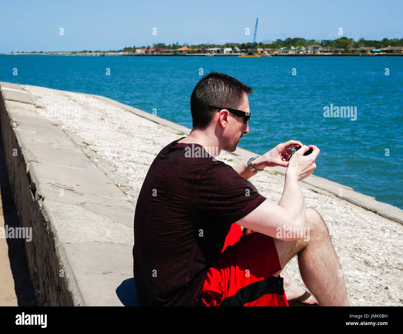 Mann mit Sonnenbrille sitzt in der Nähe der Uferpromenade mit Smartphone Stockfoto