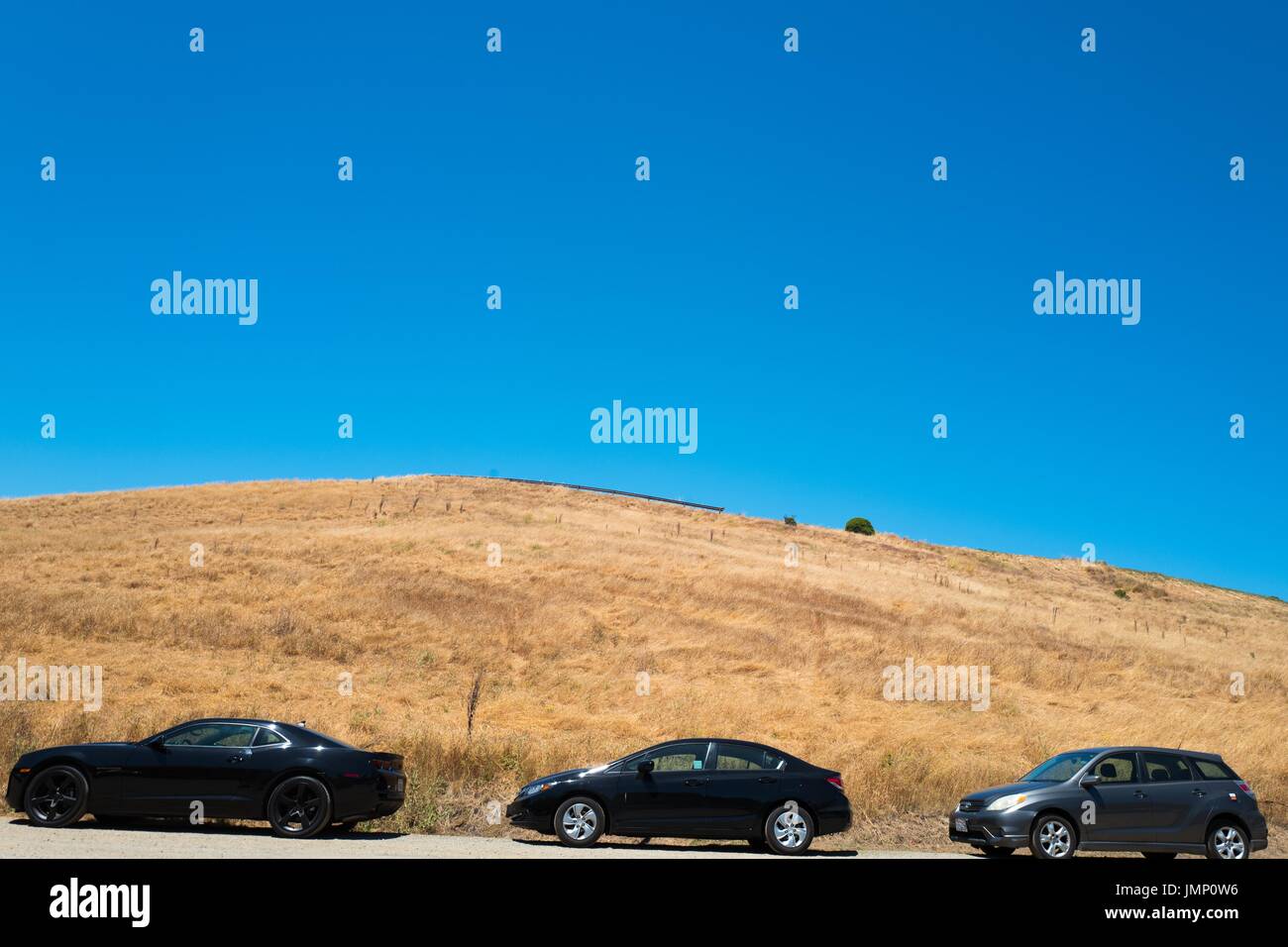 Eine Linie von drei Autos parkt in der Nähe von einem Stroh bedeckten Hügel, mit einem strahlend blauen Himmel oben, im See Chabot Regional Park, ein Regionalpark East Bay in Castro Valley, Kalifornien, 4. Juli 2017. Stockfoto