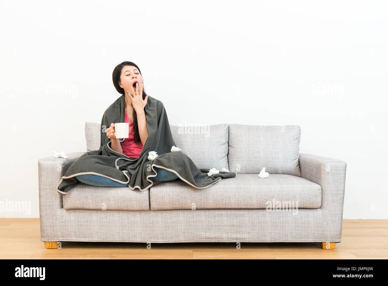 junge attraktive Frau Gefühl müde Gähnen hält heiße Tasse Becher auf Sofa sitzen, wenn sie eine Erkältung mit weißem Hintergrund in Holzboden couch. Stockfoto